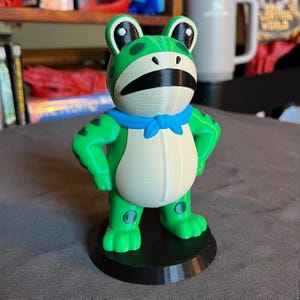 May include: A 3D-printed frog figurine. The frog has a green body, a white belly, and a blue neckerchief. It stands on a black circular base. The frog's arms are at its sides, and it has a slightly grumpy expression. The figurine is approximately 10 cm tall.