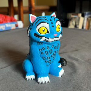 Puede incluir: Una figura azul de un gato de dibujos animados con rayas negras, manchas y dientes blancos. El gato tiene grandes ojos amarillos y el interior de las orejas rosado. La figura est&aacute; sobre una superficie gris.