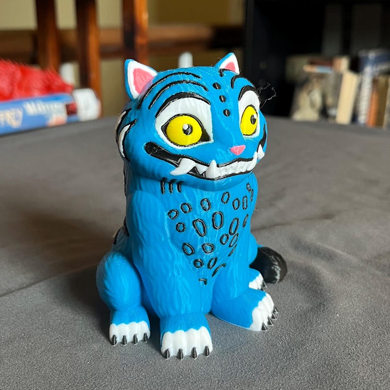 Puede incluir: Una figura azul de un gato de dibujos animados con rayas negras, manchas y dientes blancos. El gato tiene grandes ojos amarillos y el interior de las orejas rosado. La figura est&aacute; sobre una superficie gris.