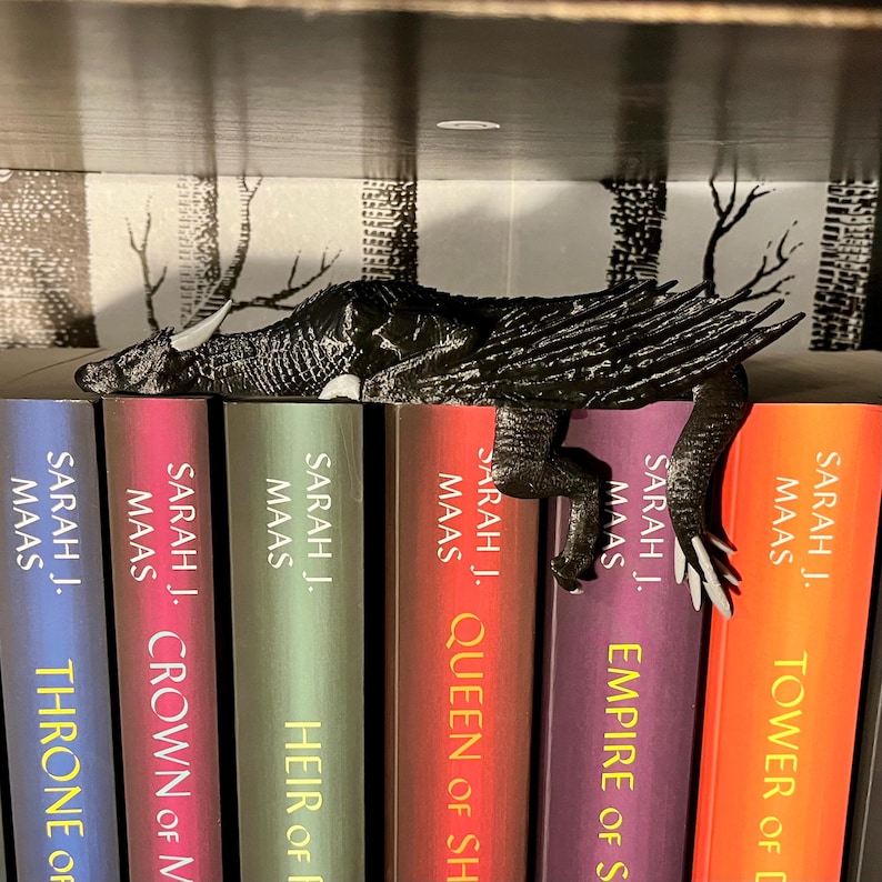 May include: A black dragon bookend resting on a bookshelf with a set of five books by Sarah J. Maas. The books are titled "Throne of Glass", "Crown of Midnight", "Heir of Fire", "Queen of Shadows", and "Empire of Storms".