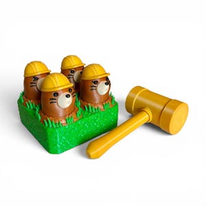 May include: A game set featuring four brown mole figures with white snouts and yellow construction hard hats, emerging from a green textured base resembling grass. A matching yellow mallet with a cylindrical head is positioned beside the base.