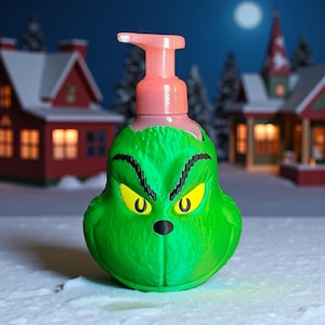 May include: A green Grinch-shaped soap dispenser with a pink pump. The dispenser features yellow eyes and a black nose. The background shows a snowy scene with houses and a full moon.