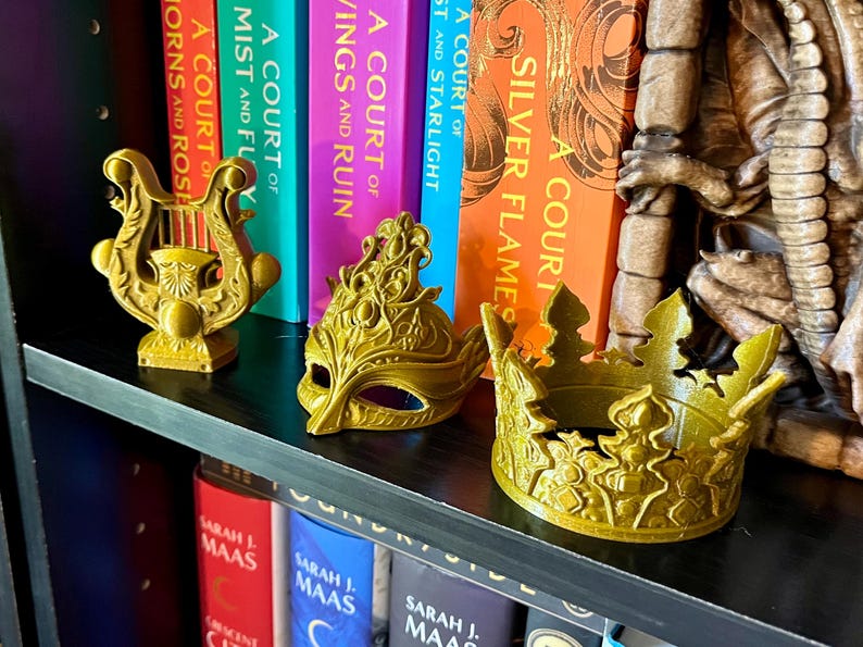 May include: Three gold-colored decorative items sit on a black shelf in front of a row of books. The items include a lyre, a masquerade mask, and a crown. The books have colorful spines with titles like "A Court of Silver Flames" and "A Court of Wings and Ruin."