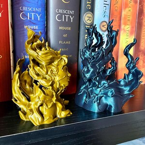 May include: Two decorative sculptures, one gold and one teal, each depicting a stylized female face with flowing, flame-like hair. The sculptures are displayed on a black shelf in front of a row of books.