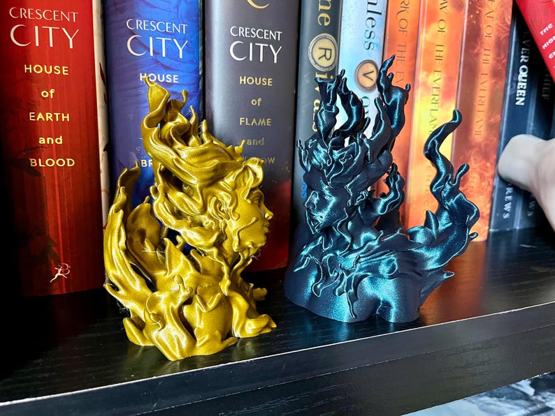 May include: Two decorative sculptures, one gold and one teal, each depicting a stylized female face with flowing, flame-like hair. The sculptures are displayed on a black shelf in front of a row of books.