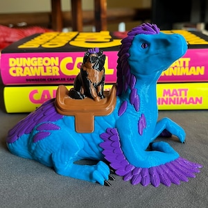 May include: A blue and purple dinosaur figurine with a small dog figurine riding on its back. The dinosaur has feathered wings and a saddle. The background includes two books with the text "Dungeon Crawler" and "Matt Inman".