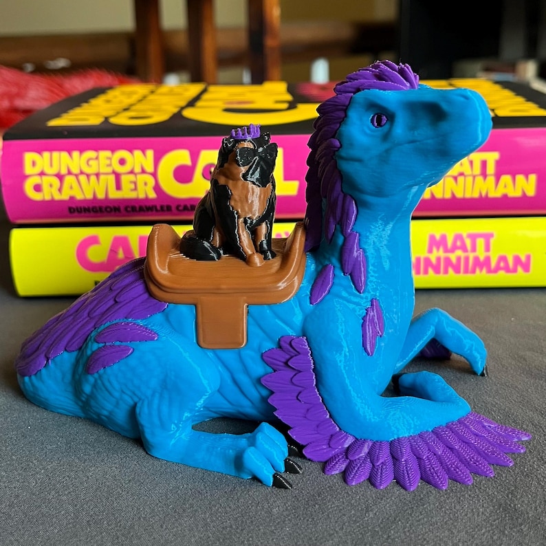 Puede incluir: Una figura de dinosaurio azul y morado con una peque&ntilde;a figura de perro montada en su espalda. El dinosaurio tiene alas emplumadas y una silla de montar. El fondo incluye dos libros con el texto "Dungeon Crawler" y "Matt Inman".