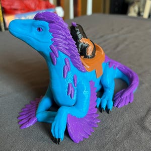 Puede incluir: Criatura de fantas&iacute;a impresa en 3D en tonos azules y morados, con una silla de montar. La criatura tiene un cuello largo, una melena de plumas y una cola curvada. La silla de montar es marr&oacute;n y negra. La criatura est&aacute; sobre una superficie gris.