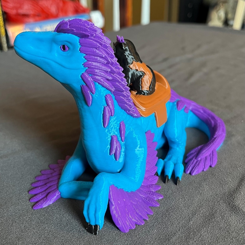 Puede incluir: Criatura de fantas&iacute;a impresa en 3D en tonos azules y morados, con una silla de montar. La criatura tiene un cuello largo, una melena de plumas y una cola curvada. La silla de montar es marr&oacute;n y negra. La criatura est&aacute; sobre una superficie gris.
