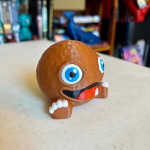 May include: A brown, spherical, 3D-printed character figurine with a textured surface. It features large, round blue eyes, a wide black mouth with a red tongue, and a single white tooth. The figure has small brown arms with white-tipped fingers.