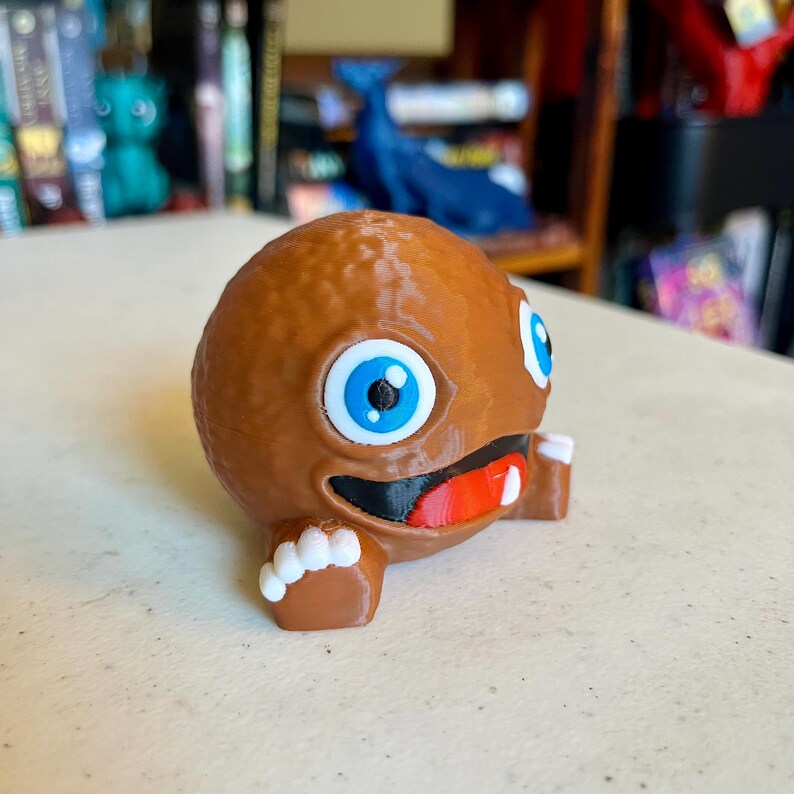 May include: A brown, spherical, 3D-printed character figurine with a textured surface. It features large, round blue eyes, a wide black mouth with a red tongue, and a single white tooth. The figure has small brown arms with white-tipped fingers.