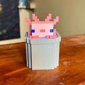 May include: A pink and blue 3D printed figurine of an axolotl in a gray block.