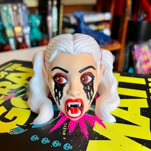 May include: A sculpted head figurine featuring white hair styled in two pigtails, pointed ears, and a pale complexion. The face has red eyes, black paint dripping from the eyes, bright red lips, and two prominent white fangs protruding from the mouth.