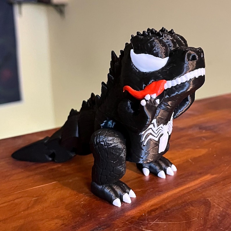 May include: A black 3D-printed Godzilla figure with white teeth, eyes, and a red tongue. The figure has a Venom-inspired design, including the white spider symbol on its chest.
