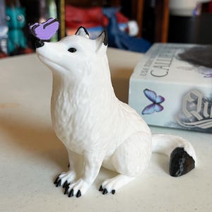 May include: A white figurine of a fox with black accents on its ears and paws, gazing upwards at a purple butterfly perched on its nose. The fox is sitting, with its tail curled. A box with a butterfly graphic is in the background.