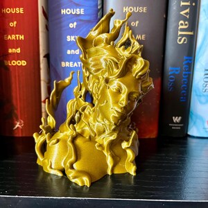 May include: A gold-coloured sculpture of a woman's bust, with flowing, flame-like details. The sculpture is positioned in front of a shelf with books, including titles like "House of Earth and Blood" and "Rivals".