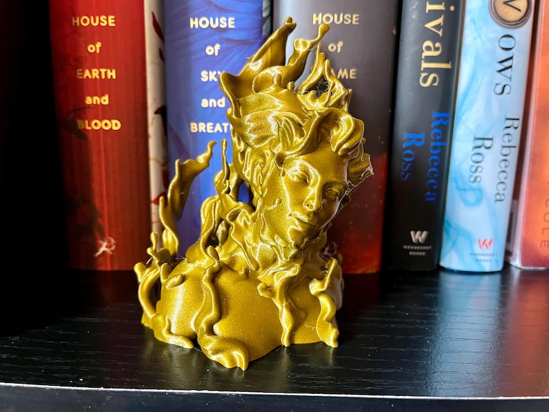 May include: A gold-coloured sculpture of a woman's bust, with flowing, flame-like details. The sculpture is positioned in front of a shelf with books, including titles like "House of Earth and Blood" and "Rivals".