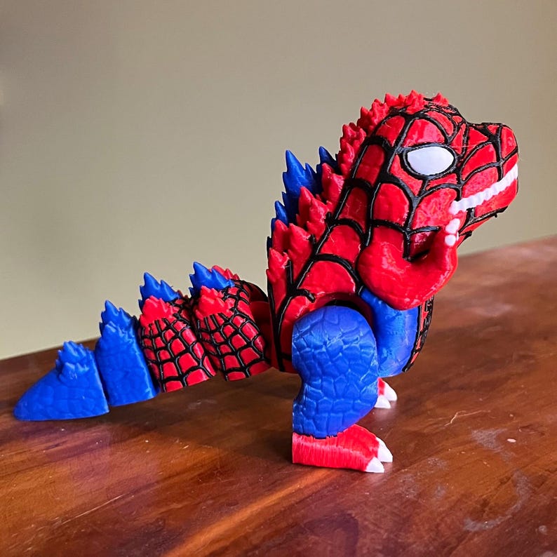 May include: A 3D-printed toy dinosaur in red, blue, and black. The dinosaur has a red head with a white eye and a web pattern, blue legs, and a blue and red tail. The toy is on a wooden surface.