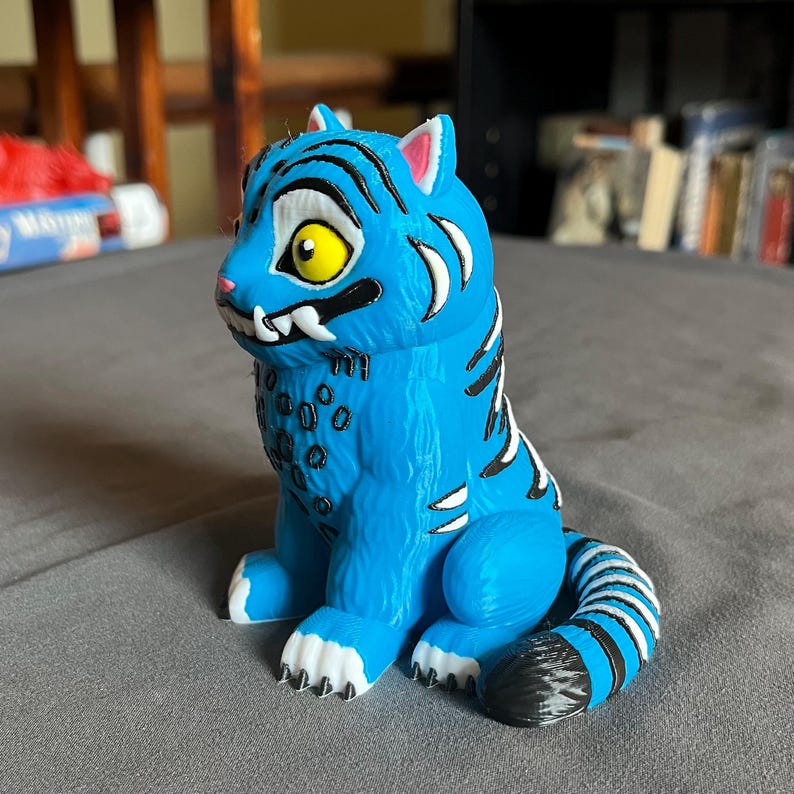 Puede incluir: Una figura de tigre azul con rayas negras y detalles blancos. El tigre tiene un ojo amarillo, dientes blancos y una cola a rayas blancas y negras. La figura est&aacute; sobre una superficie gris.