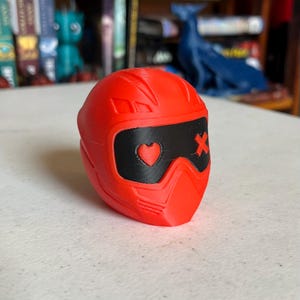 K&ouml;nnte beinhalten: Ein roter, helmformiger Gegenstand mit schwarzem Visier. Das Visier zeigt ein Herz und ein X-Symbol. Der Gegenstand ist wahrscheinlich ein Dekorationsartikel oder Spielzeug, m&ouml;glicherweise 3D-gedruckt.
