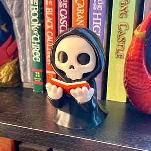 May include: A black and white figurine of a Grim Reaper reading a red book. The figurine has a skull face and is wearing a black hooded robe. The figurine is on a shelf in front of a row of books.
