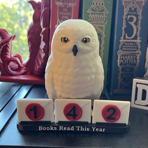 Puede incluir: Una figura de búho blanco con ojos amarillos se encuentra sobre una pantalla que dice "Books Read This Year". La pantalla muestra los números 1, 4 y 2 en pequeños bloques con forma de sobre. El búho está frente a una estantería con libros.
