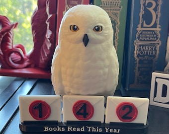 Op de afbeelding: Een witte uilfiguur met gele ogen zit boven een display met de tekst "Books Read This Year". De display toont de nummers 1, 4 en 2 op kleine envelopvormige blokken. De uil staat voor een plank met boeken.