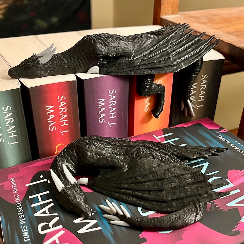 May include: Two black dragon bookends with white claws, resting on a stack of books with the author's name "SARAH J. MAAS" printed on the spines. The top book is titled "MIDNIGHT", with the author's name "SARAH J. MAAS" printed on the spine.