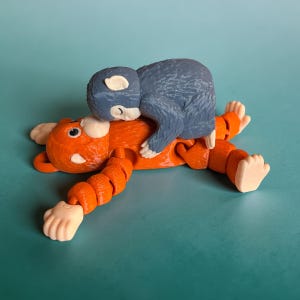 May include: A whimsical orange and blue figurine set. The orange figure is lying down with its arms and legs outstretched. A blue figure is on top of the orange figure. The figures are on a teal background.