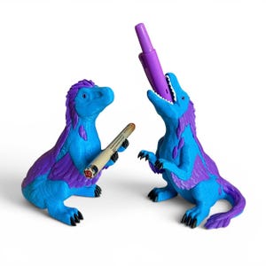 May include: Two blue and purple dinosaur-shaped pen holders. The left dinosaur holds a thin beige pen with a red cap, while the right dinosaur has its mouth open, holding a purple pen vertically. Both figures have textured skin and black claws.
