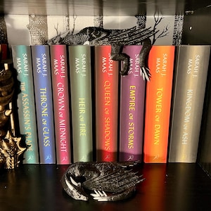 May include: A bookshelf with a black dragon bookend. The bookshelf is filled with fantasy novels by Sarah J. Maas. The titles of the books are "The Assassin's Blade", "Throne of Glass", "Crown of Midnight", "Heir of Fire", "Queen of Shadows", "Empire of Storms", "Tower of Dawn", and "Kingdom of Ash".