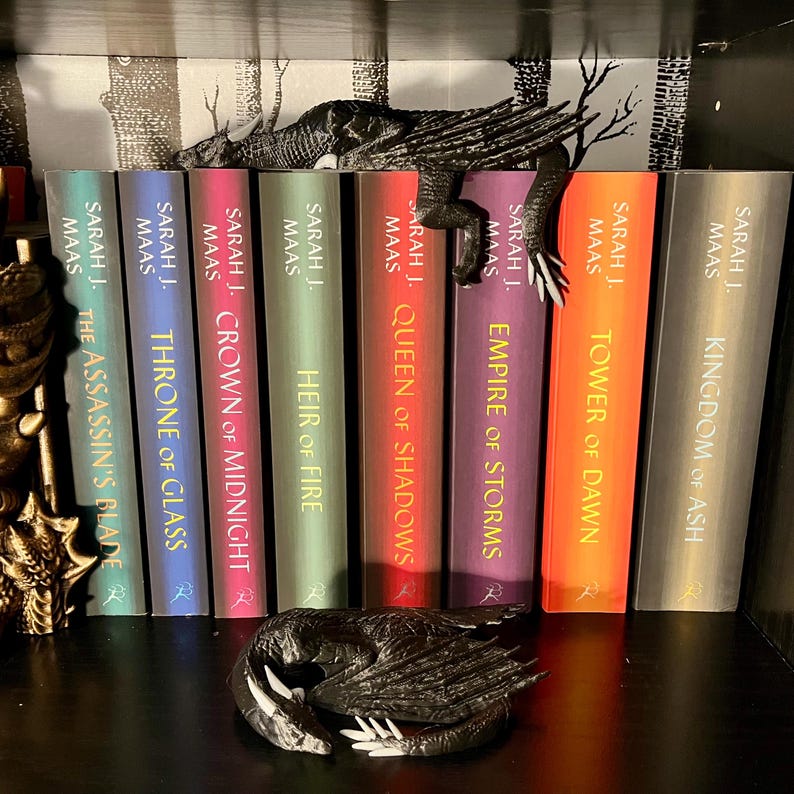 May include: A bookshelf with a black dragon bookend. The bookshelf is filled with fantasy novels by Sarah J. Maas. The titles of the books are "The Assassin's Blade", "Throne of Glass", "Crown of Midnight", "Heir of Fire", "Queen of Shadows", "Empire of Storms", "Tower of Dawn", and "Kingdom of Ash".
