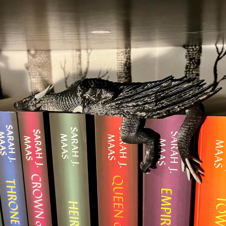 May include: A black dragon bookend resting on a bookshelf with a row of books by Sarah J. Maas. The books are titled "Throne of Glass", "Crown of Midnight", "Heir of Fire", "Queen of Shadows", "Empire of Storms", and "Tower of Dawn".