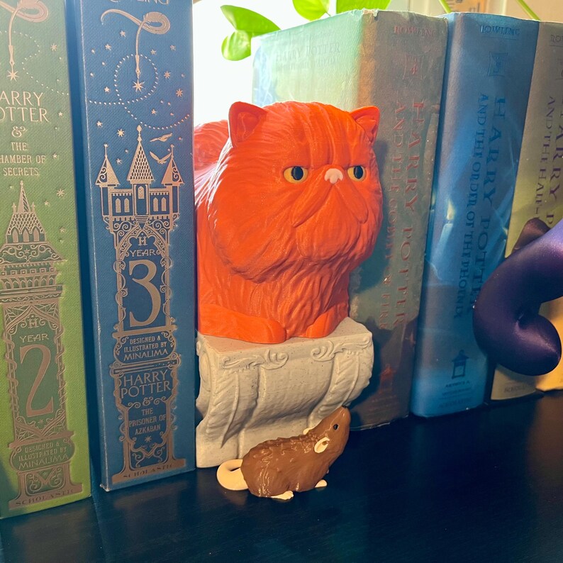 Może przedstawiać: Pomarańczowa figurka kota stoi między książkami o Harrym Potterze. Mała brązowa figurka myszy znajduje się przed kotem. Książki są w r&oacute;żnych kolorach, w tym zielonym i niebieskim. Kot stoi na ozdobnym cokole.