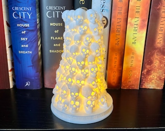 May include: A white ceramic skull candle holder shaped like a tree. The candle holder is lit and glowing, casting a warm light. The candle holder is sitting on a shelf with several books behind it.