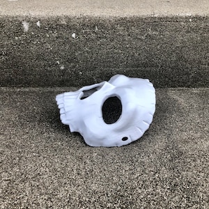 Peut inclure: Un masque de cr&acirc;ne blanc, couvrant la moiti&eacute; inf&eacute;rieure du visage, avec des ouvertures pour les yeux et la bouche. Le masque est fait d'un mat&eacute;riau lisse. Il repose sur une surface grise textur&eacute;e. Le masque est un costume d'Halloween.