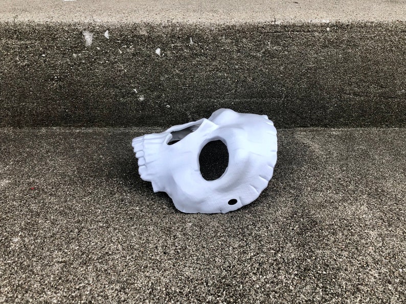 Peut inclure: Un masque de cr&acirc;ne blanc, couvrant la moiti&eacute; inf&eacute;rieure du visage, avec des ouvertures pour les yeux et la bouche. Le masque est fait d'un mat&eacute;riau lisse. Il repose sur une surface grise textur&eacute;e. Le masque est un costume d'Halloween.