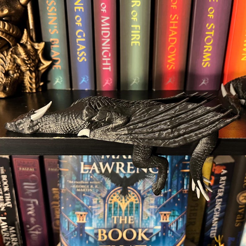 May include: A black dragon bookend with white claws, perched on a bookshelf with a row of fantasy novels. The dragon is resting its head on the shelf, with its wings spread out.