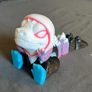 Könnte beinhalten: Eine 3D-gedruckte Figur einer dinosaurierartigen Kreatur in sitzender Position. Die Figur hat einen weißen Kopf mit rosa und roten Akzenten, blaue Hände und Füße sowie einen schwarzen und blauen Körper. Der Schwanz ist schwarz und blau.