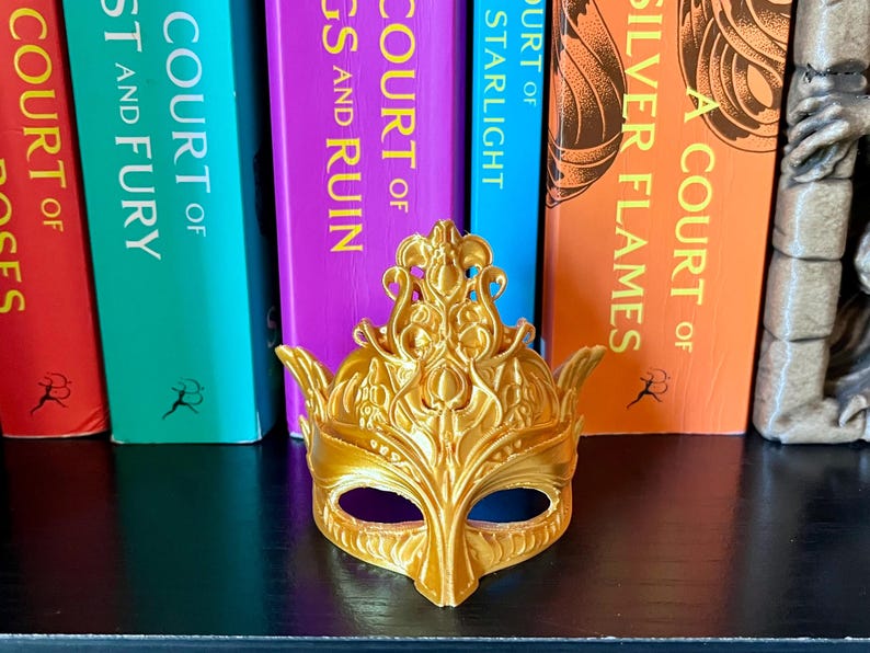 May include: A gold-colored decorative mask with an ornate design, featuring a crown-like top and eye openings. The mask is positioned in front of a row of colorful books. The books have titles such as "A Court of Silver Flames".