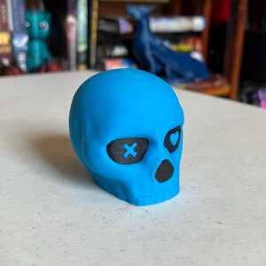 K&ouml;nnte beinhalten: Eine hellblaue Totenkopf-Figur mit schwarzen Augenh&ouml;hlen und Nasenh&ouml;hle. Eine Augenh&ouml;hle hat ein X, die andere ein Herz. Der Sch&auml;del liegt auf einer wei&szlig;en Oberfl&auml;che.