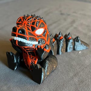 Könnte beinhalten: Eine 3D-gedruckte, stilisierte Dinosaurierfigur in Schwarz und Orange. Der Dinosaurier hat ein detailliertes, netzartiges Muster auf Kopf und Brust, mit einem einzelnen weißen Auge. Sein stacheliger Rücken und Schwanz sind ebenfalls sichtbar.