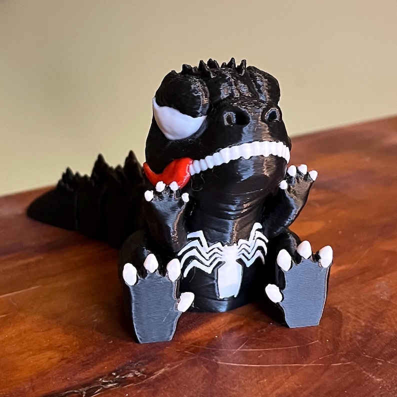 May include: A black and white 3D-printed figurine of a cartoonish dinosaur with a red tongue sticking out. The figurine has white teeth, claws, and eye details. The chest features a white spider-like emblem.