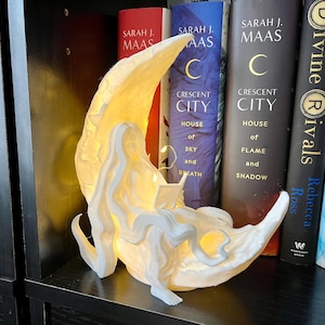 May include: A white 3D printed lamp shaped like a crescent moon with a woman sitting on it reading a book. The lamp is lit up with warm white light. The lamp is sitting on a bookshelf with several books behind it.