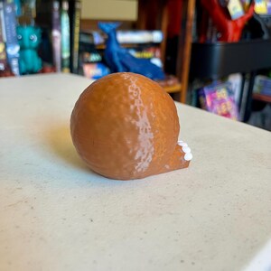 May include: A brown, spherical, 3D-printed object with a textured surface. A small, protruding base section is visible on the right side, featuring three distinct, small, white, circular accents arranged in a horizontal row on its edge.