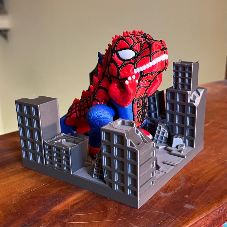 May include: A red, blue, and white Spider-Man-themed Godzilla figurine destroying a miniature city. The figurine has a detailed design, with a red and black web pattern on its head and body. The city is gray, with several buildings in various states of destruction.