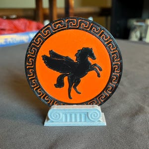 May include: A 3D-printed decorative item featuring a black Pegasus silhouette on an orange background. The circular design has a black border with a repeating pattern. The base is a light blue, column-style support.