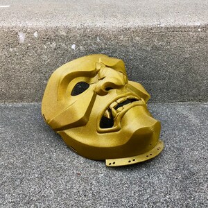 May include: A gold-coloured, detailed, plastic mask with a fierce expression. The mask has a wide mouth with sharp teeth and a prominent nose.