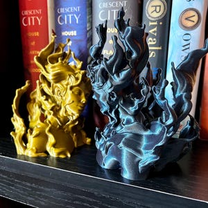 May include: Two sculptures of a woman's face with flowing hair, one gold and one dark blue. The sculptures are in front of a bookshelf with books that have the text "CRESCENT CITY" and "THE EVERFLAME".