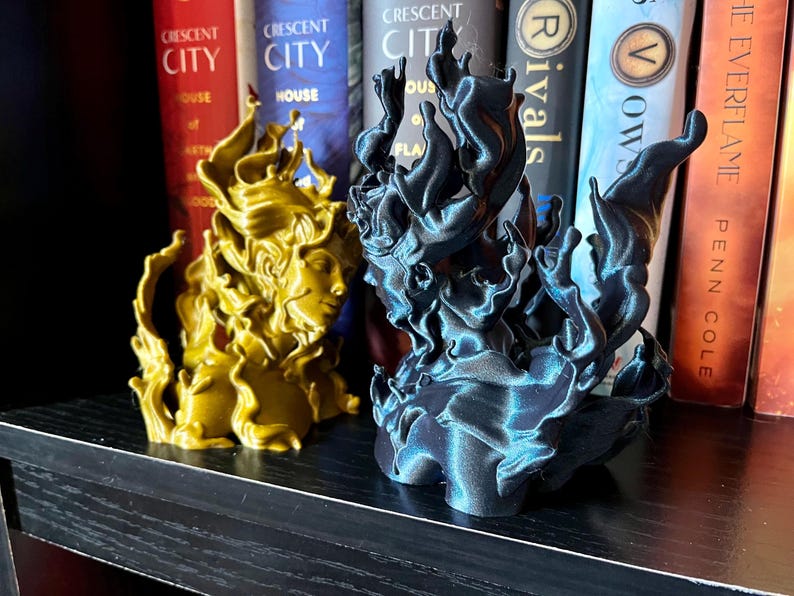 May include: Two sculptures of a woman's face with flowing hair, one gold and one dark blue. The sculptures are in front of a bookshelf with books that have the text "CRESCENT CITY" and "THE EVERFLAME".